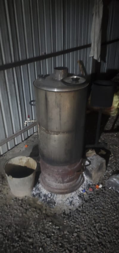 In Abdurahmoni Jomi, Tajikistan, a large, cylindrical metal boiler is positioned outdoors, resting on a base of ash and embers. The boiler appears to be made of stainless steel and has two handles on its sides. A metal spigot with a red handle is attached to the side of the boiler, near its base. The boiler is placed on a surface covered with dark gravel and ash. To the left of the boiler is a light-colored metal bucket. Behind the boiler is a corrugated metal wall. To the right of the boiler, a dark metal stand supports a black container, and some yellow material is visible behind it. A piece of white cloth is hanging on the corrugated metal wall. Several small, colorful plastic caps are scattered on the gravel in front of the boiler's base. The scene appears to be an outdoor cooking or heating setup.