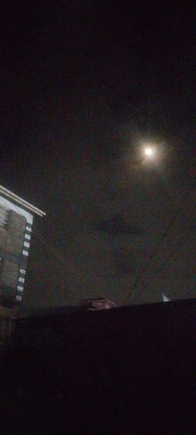 The image is a night scene captured in Ndenderu ward, Kenya. A bright, full moon is visible in the dark, cloudy sky. In the lower left portion of the image, the corner of a building with white trim and horizontal striped windows is visible. In the foreground, a dark, raised surface obscures the lower part of the image, with a small, indistinct object on its surface. Electrical wires are also visible crisscrossing the scene. The overall impression is one of a nocturnal urban environment.