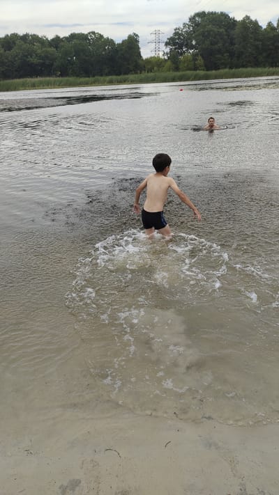 The image depicts a shallow body of water, possibly a lake or river, with a sandy bottom visible near the foreground. A young boy, wearing black swim trunks, is standing in the water, creating splashes with his movement. His back is turned to the camera, and his arms are slightly extended to the sides. Further in the water, a man is swimming, with his head and shoulders visible above the surface. The water is calm, with ripples and some foam generated by the boy's activity. In the background, a line of lush green trees forms the horizon, with a cloudy sky overhead. A tall electrical pylon is visible amongst the trees. The overall scene suggests an outdoor recreational setting, likely a beach or a waterfront park. The location is identified as Ramon, Russia, indicating it is a natural or semi-natural area within or near the city.