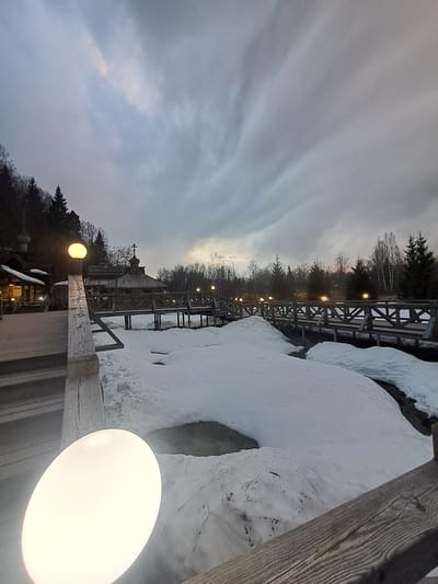 The image depicts a landscape in Vzglyadnevo, Russia, featuring a wooden boardwalk or bridge covered in snow. The scene is dominated by overcast skies. A traditional Russian wooden church with a cross on top is visible in the background, partly obscured by trees. The boardwalk is lined with illuminated lampposts. Snow covers the ground on either side of the boardwalk. A portion of a wooden railing or structure is prominently featured in the foreground, partially obscuring a lamppost.