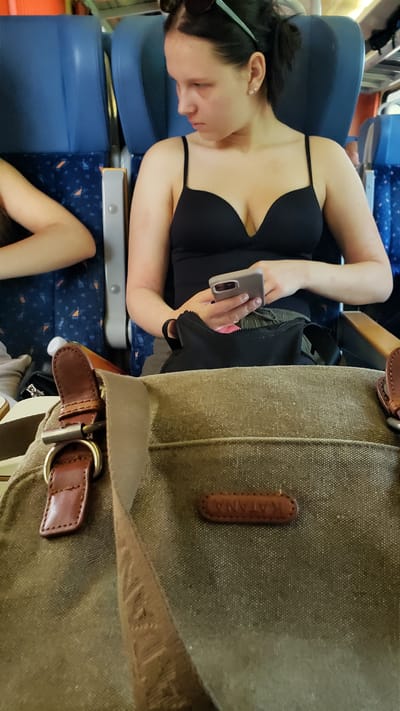 The image shows a young woman sitting on a train. She is wearing a black top and looking at her phone. In the foreground, there is a close-up of a brown canvas bag with leather accents. The scene appears to be inside a train carriage, indicated by the blue seats and the window view. The location is Púchov, Slovakia.