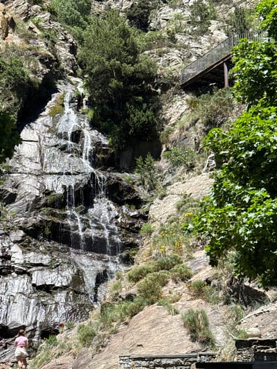 The image depicts a steep, rocky mountainside with a cascading waterfall. A metal footbridge, likely part of a hiking trail, is visible on the upper left, leading over a crevice. Lush green foliage, including trees and smaller plants, grows on the rocks, especially in the shaded areas. The waterfall tumbles down a dark, wet rock face, creating streaks of white water that sparkle in the sunlight. In the lower right, a person wearing a pink shirt and white shorts is standing, observing the waterfall. The scene appears to be set during the daytime, with bright sunlight illuminating the rocks and water. The overall impression is one of a natural, rugged landscape, possibly a popular spot for outdoor recreation.