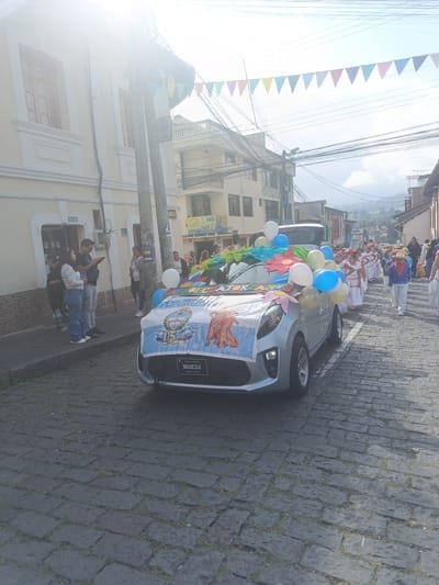 The image captures a daytime scene on a cobblestone street in Amaguaña, Ecuador, during what appears to be a celebratory procession. A light-colored compact car, possibly silver or light gray, is positioned centrally in the foreground, facing away from the viewer. The vehicle is elaborately decorated: a large banner across its front displays the Argentine flag design, a "15 AÑOS" emblem, an illustration of a woman in traditional dress, and partially visible text including "PRODUCTO ECUATORIANO" and "AMARTE DESDE LA SALUD." The car's hood and front windshield area are covered with green leafy garlands, colorful paper flowers (red, pink, orange), and an array of white, blue, and yellow balloons. A "Sparco" logo is visible below the banner, near the license plate area.
On the left side of the street, three adults are standing on a paved sidewalk. A woman in a patterned white long-sleeved shirt and blue jeans stands next to a man in a black t-shirt and light gray pants; both are looking down, possibly at mobile phones. Further back, another woman in a gray long-sleeved top stands near a pale yellow building with white trim and brown wooden windows. A utility pole with numerous black power lines and a street sign displaying "Qst. 18" and "VIA" is prominently located on this sidewalk.
To the right of the decorated car, a line of people, primarily children, walks down the street in what appears to be a parade. Many of the children are dressed in traditional festive attire, consisting of white dresses with red sashes and details, some wearing straw hats. A boy near the front of this procession wears white pants, a blue top with a red sash, and a straw hat. An adult male in a dark jacket follows the group further down the street.
Overhead, multiple black power lines crisscross the sky. A string of colorful triangular bunting flags (red, orange, yellow, green, blue, purple) is strung across the street, indicating a festive atmosphere. In the distant background, green hills or mountains are visible under a partly cloudy sky. The cobblestone street features some minor vegetation growth between the stones. Bright sunlight from the upper left quadrant causes a visible lens flare in the image.