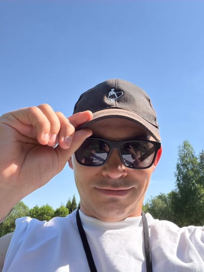 The image depicts a person in Oreshniki, Russia, wearing a white t-shirt, a dark-colored baseball cap with a small white emblem on the front, and sunglasses. The person is holding the brim of the cap with their hand. The background consists of a clear blue sky and some green trees, suggesting an outdoor location. The person appears to be taking a selfie, based on the camera angle and the positioning of their hand. A lanyard hangs around the person's neck.