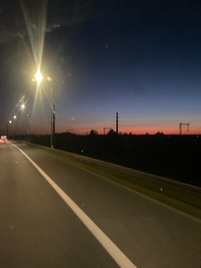 This media file depicts a dusk or dawn scene from the perspective of someone traveling along a road, likely a highway, in Węgrzce Wielkie, Poland. The sky transitions from a deep blue at the top to bands of pink, orange, and lighter blue near the horizon, indicating twilight. Silhouetted against the colorful sky are power lines and trees, suggesting a rural or semi-rural landscape.
The foreground is dominated by the asphalt road, marked with a white line on the right side. The road surface appears wet, reflecting the ambient light. On the left side of the frame, a bright streetlight emits a strong glow, creating a lens flare effect and illuminating the surrounding area. Other smaller streetlights are visible further down the road. The overall impression is one of travel at a transitional time of day, with artificial lights beginning to dominate the scene as natural light fades or emerges. There is no visible text in the image. The mood is serene and perhaps a bit melancholic due to the dim light and solitary nature of the scene.