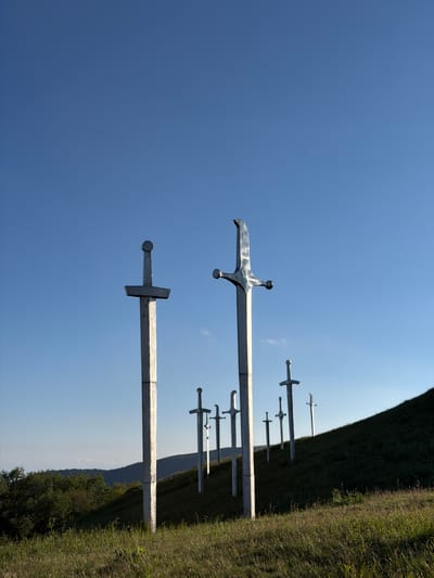 This media file features a series of large, metallic sword sculptures planted vertically in the ground. The swords are silver in color and appear to be made of metal. They are arranged in a line, receding into the background, with some of them partially obscured by a grassy hill. The scene is set against a clear, bright blue sky, suggesting a sunny day. The perspective is from a low angle, looking upwards at the swords and the sky. The ground is covered in green grass, and a distant hillside is visible in the background, partially covered with trees. The overall impression is one of a monument or an outdoor art installation. The time of day appears to be daytime, likely midday or afternoon, given the brightness of the sky. There is no visible text in the image. The location context provided is Zemo Nichbisi, Georgia.