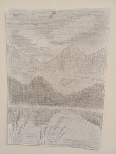 This is a pencil sketch of a serene, mountainous landscape at dusk or dawn. The scene is dominated by layered mountain ranges, rendered with soft, hazy pencil strokes, suggesting distance and atmospheric perspective. In the sky, a pale circle, likely the moon or a dim sun, is visible, further contributing to the subdued lighting. Below the mountains, a body of water, possibly a lake or river, reflects the dim light and the silhouettes of the land. The foreground features a gentle slope leading to the water's edge, with wisps of tall grass or reeds sketched along the bank. The overall mood is peaceful and tranquil. There is no visible text in the image. The sketch is pinned to a wall with a white pushpin.