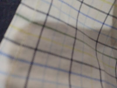 The image displays a close-up view of a checkered fabric, likely a shirt or a tablecloth. The pattern consists of thin, intersecting lines of blue, yellow, and dark gray on a white background. The fabric appears to be slightly creased, suggesting it is either worn or loosely laid out. The lighting is even, with no strong shadows. The image is out of focus, making it difficult to discern specific details of the fabric's texture or any additional objects that might be present. There are no people or discernible human activity depicted. The background to the left of the fabric is a dark, out-of-focus area that appears to be a solid color, possibly a piece of clothing or upholstery. There is no information within the image to indicate a location in Eliowhani, Nigeria.