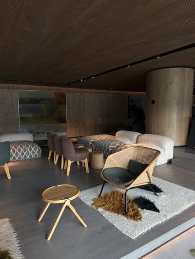 An overhead shot captures a modern, upscale lounge or waiting area, characterized by a sophisticated and warm aesthetic. The dark wood plank flooring runs throughout the space, providing a rich foundation. A prominent dark brown perforated wall, accented with sleek black strips and small embedded lights, stretches across the left side of the frame, curving slightly upwards. In the background, a lighter wood panel wall is visible, complementing the dark flooring.
The lounge is furnished with a variety of contemporary pieces. A large, light-colored textured sofa or bench, possibly modular, is adorned with a woven throw blanket. Adjacent to it, a stylish armchair crafted from light wood and cane webbing, featuring a dark grey cushion, stands out. Underneath this chair lies a shaggy, off-white rug with fringed edges, on which two decorative, shaggy elements—one black and one brown—resemble abstract shapes or animal features.
Further into the room, two dark brown upholstered armchairs with light wooden legs are arranged, positioned near a large, multi-panel framed artwork on the wall. This artwork displays a series of serene landscape or seascape paintings in muted tones. A small, round wooden side table with three splayed legs is placed on the dark flooring, with a tiny, illegible tag visible on its surface. Another small, textured, round stool is also present. The overall lighting appears bright, suggesting a daytime setting or a well-lit interior, creating a tranquil and inviting atmosphere. No people are visible.