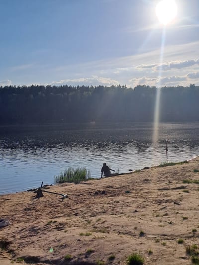 The scene is a bright day at a lake in Меткомелино, Russia. A sandy beach borders the water. A person is standing in the shallow water near the beach. Further out in the lake, a thin post rises from the water. The opposite shore is lined with dense trees. The sky is blue with white clouds and the sun is shining brightly. An anchor is on the beach.
