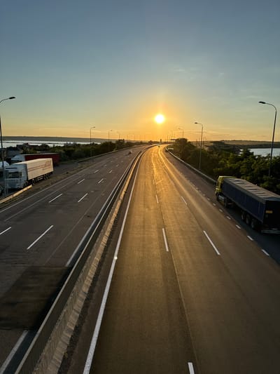 The image shows a highway in Yelyzavetivka, Ukraine, during sunset. The sun is visible in the sky, casting a warm glow on the road and the surrounding landscape. The highway has multiple lanes and is bordered by a concrete barrier and streetlights. Several trucks are parked on the side of the road, and one truck is driving on the highway. In the distance, there is a body of water and some trees. The sky is clear with a gradient of blue and orange colors.