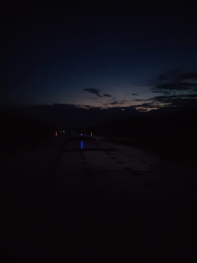 The image depicts a night scene in Aleksandrovskoye, Russia. A road or walkway is visible in the foreground, appearing dark with some lighter-toned patches that suggest concrete or a similar material. There are footprints visible on this surface. In the midground, there are scattered lights, some with a blue hue and others with a reddish hue, likely from streetlights or other artificial sources. The background shows a dark sky with some cloud cover. There is a hint of twilight or the very beginning of sunrise on the horizon, with faint colors peeking through the clouds. Two faint stars are visible in the upper portion of the sky. The overall scene is dim and obscured by darkness.