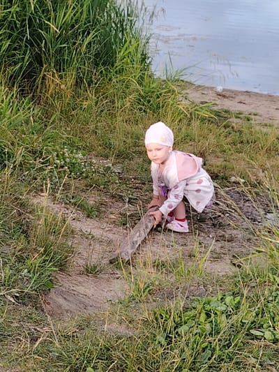 A young girl, wearing a pink headscarf and a white dress with pink polka dots, squats on a grassy embankment, holding a piece of wood. She is positioned in the foreground of the image, with tall green grass surrounding her. In the background, a body of water is visible, with a sandy shore. The scene appears to be a natural outdoor setting, possibly a park or the edge of a lake. The location is identified as Zayamanauki rural council, Belarus.