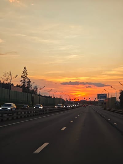 The image captures a highway scene at sunset, likely in or near посёлок опытного хозяйства «Ермолино», Russia. The sky is ablaze with vibrant orange and yellow hues, indicative of a dramatic sunset, with scattered clouds adding texture. A multi-lane highway stretches into the distance, bordered by sound barriers on the left and right. Numerous cars are visible on the highway, most in motion and appearing as streaks of light due to the long exposure or motion blur. Streetlights line the highway, and trees and foliage are visible beyond the sound barriers, suggesting a suburban or semi-rural setting. Traffic signs are present along the roadside. The perspective is from a vehicle moving along the highway.