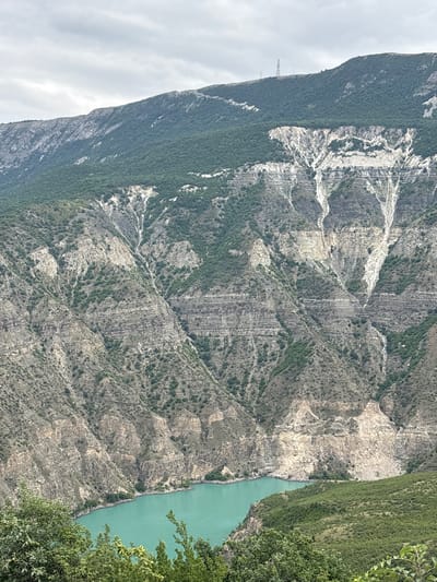 The image depicts a dramatic mountain landscape with a striking turquoise body of water at its base. The mountains are characterized by steep, layered rock formations, with streaks of lighter material, possibly scree or mineral deposits, running down their faces. Vegetation, ranging from sparse shrubs to denser green foliage, clings to the slopes and covers the higher mountain peaks. In the distance, on the crest of one of the highest mountains, a tall, lattice-like transmission tower is visible. The sky is overcast with soft, gray clouds. The foreground is partially obscured by the leaves of green trees. This scene is located near the village of Zubutli, Russia, likely showing a gorge or canyon feeding into a reservoir or a large river.