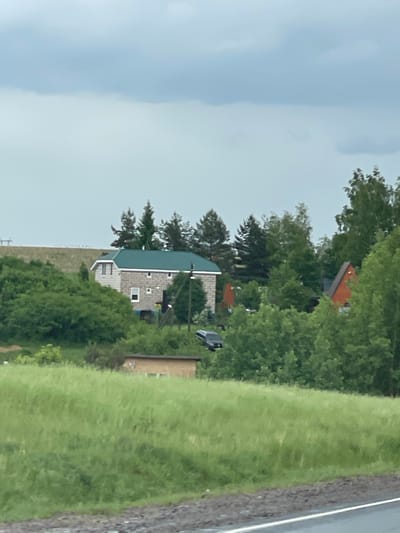 The image shows a rural scene in Кокино, Russia. A light-colored house with a green roof sits amidst lush greenery. A car is parked nearby. To the right of the house is a field of grass. The sky is overcast. Trees are visible in the background. A road is to the right of the frame.