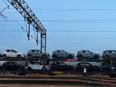 The image depicts a train car carrier filled with cars, likely for transportation. The train is located near or within the city of Ruma Municipality, Serbia, indicated by the "TRANSWAGGON" and "BERTANI" branding, both of which are associated with logistics and transportation companies operating in Europe. The train cars are carrying multiple levels of vehicles, primarily small SUVs or crossovers. The color scheme includes white, black, and gray cars. A person is walking on the ground near the train. An overhead electrical grid is visible above the train, possibly for powering electric locomotives. The sky is pale blue.