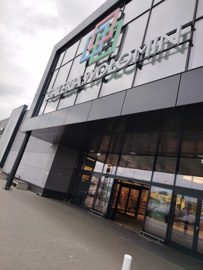 The image shows the exterior of a modern shopping center, identified by the signage as "GALERIA WOŁOMIN". The building features a prominent glass facade with the shopping center's name displayed in large, white, three-dimensional letters. Above the name, a colorful logo composed of interlocking geometric shapes is visible. The entrance to the shopping center consists of large glass doors, which are open, revealing the interior with bookshelves lining the walls. The pavement in front of the entrance is made of interlocking concrete pavers. To the left of the main entrance, a large advertisement poster is displayed vertically. The sky above is overcast. The perspective suggests the photo was taken from ground level, looking upwards towards the building.
