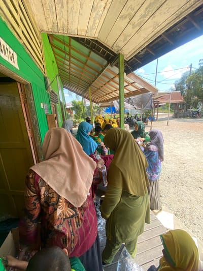 In Banyu Hirang, Indonesia, a group of people are gathered outdoors under a shaded wooden structure, likely part of a building's overhang or a covered walkway. The building to the left is painted bright green, with a sign that reads "KANTOR" (office) in white letters. The ground is covered in gravel. Many individuals, including children and adults, are wearing hijabs, with several adults in the foreground wearing predominantly green and beige headscarves. Some are interacting with what appears to be a distribution of items, with hands reaching for bags or packages. Several motorbikes are parked in the background, suggesting a rural or semi-rural setting. The sky is visible with some clouds. The overall scene appears to be a community gathering or event involving the distribution of goods.