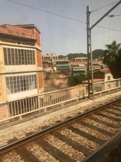 The image captures a view from a moving train in Montcada i Reixac, Spain. The foreground shows train tracks with ballast and wooden sleepers. In the midground, a bridge railing separates the tracks from a built-up area. To the left, a building with a yellow lower facade and an orange upper facade features large windows with white frames. Behind this, several other buildings are visible, some with balconies and awnings. A tall, grey utility pole with overhead wires stands prominently to the right of the bridge. Further in the background, green hills and a palm tree are discernible against a hazy sky.