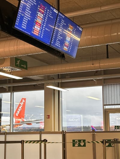 An airport departure screen displays flight information, including destinations such as Warsaw, Barcelona, Skopje, and Vienna. Some flights are marked as "Closed," and airlines like easyJet are listed. An easyJet airplane is visible through a window. A sign reads "Welcome to the CIP lounge". An exit sign is also present. The airport appears to be in Cardiff, with "DFF" displayed on a sign, possibly referring to Cardiff Airport.