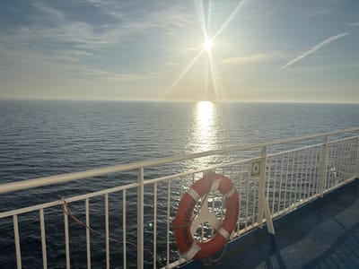 A wide-angle, medium shot captures a serene ocean scene from the deck of a ferry. The sun, positioned slightly left of center in the upper portion of the frame, emits bright rays and casts a shimmering reflection on the calm, deep blue water. A thin contrail stretches across the pale blue sky. In the foreground, a white metal railing lines the edge of the ferry deck, with a bright orange life preserver attached to it on the right side. The life preserver has white lettering that reads "ROBIN HOOD" and "EMDEN". The blue deck of the ferry is visible in the lower right corner. The scene is devoid of people, emphasizing the vastness of the sea and the sky.