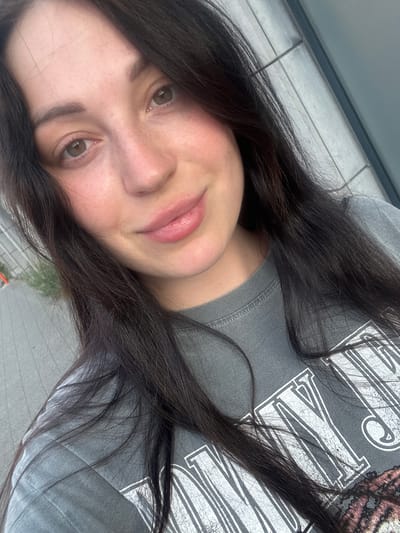 A close-up, selfie-style photograph features a young woman with long, dark brown hair and light brown eyes, looking directly at the camera with a slight smile. She is wearing a dark gray t-shirt with a graphic print. The background appears to be an outdoor urban setting with a textured, light-colored wall and some greenery visible on the left. The lighting suggests it is daytime. The image appears to have been taken in Strombeek-Bever, Belgium.