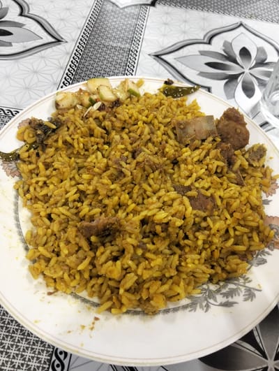 A plate of kacchi biriyani sits on a patterned tablecloth. The biriyani contains rice, chunks of meat, green chilies, and a small portion of what appears to be cucumber salad. The plate has a floral design around the rim. The scene is likely in Naodoba, Bangladesh. A glass is partially visible in the upper right corner.