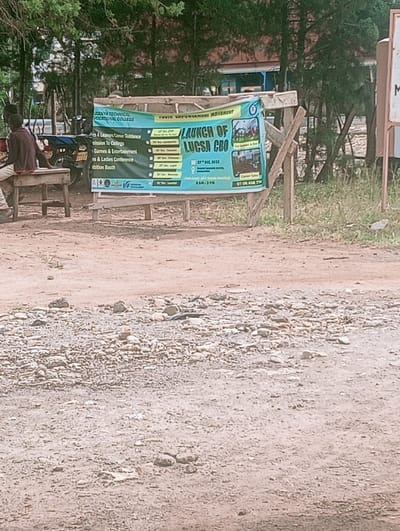 The scene depicts an outdoor environment in Lwandeti ward, Kenya. In the foreground, the ground consists of reddish-brown dirt mixed with numerous small stones and gravel. A large green and blue banner, mounted on a rustic wooden frame, dominates the midground. The banner advertises the "LAUNCH OF LUGSA CBO" by the "TWIN EMPOWERMENT MOVEMENT" and "LUGENYA TECHNICAL VOCATIONAL COLLEGE," listing event details for December 2019, including career guidance, games, a ladies conference, and an exhibition booth, along with contact information. To the left of the banner, a single individual is seated on a light-colored wooden bench, facing away from the viewer, wearing dark clothing and light-colored trousers. Two motorcycles are partially visible behind this individual and to the left of the banner. The background features dense green foliage and trees, with a building exhibiting a blue roof and reddish-brown walls partially visible through the vegetation. A white signpost with dark lettering is visible on the far right edge of the image.