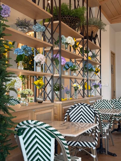 This image shows a well-lit interior scene, likely a cafe or restaurant, with a focus on a shelving unit filled with decorative floral arrangements. The shelves are made of light-colored wood and supported by a dark metal frame with an open lattice design. Various flowers, including hydrangeas in shades of blue, pink, and purple, as well as yellow and white flowers, are displayed in glass vases. Some shelves also hold greenery in woven baskets.
In the foreground, there are tables with marble-patterned tops and chairs with a distinctive black and white chevron pattern on their woven backs and seats. A tall evergreen tree or shrub is visible on the left side of the frame. The overall ambiance suggests a bright, possibly European-inspired, indoor dining or retail space. There are no people visible in the image. The location is identified as Ban Samnak Tabaek, Thailand.