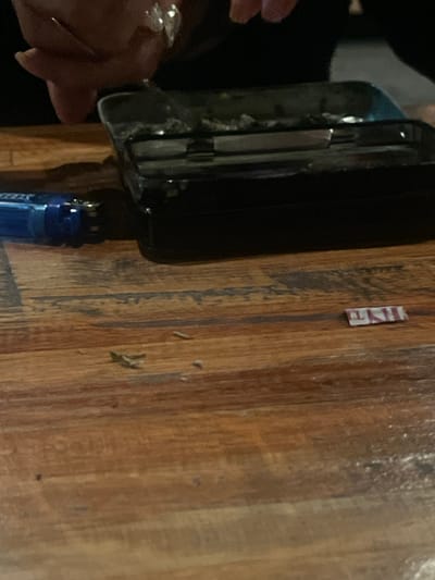 A black smartphone is partially held by a hand with visible rings on a wooden surface. An item with "MINE" printed on it is on the surface. A blue bottle-like object is partially visible attached to the smartphone. The location and any interactions cannot be determined from the image.