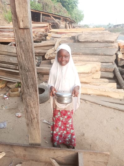 A young girl, likely of Muslim faith, stands smiling in a cluttered outdoor area. She wears a white hijab and a vibrant red and blue patterned skirt. She holds a metal container with handles. Behind her, stacks of lumber are visible, and a weathered wooden structure is in the background. The scene seems to be a working area, possibly a sawmill, with a mix of wood, dirt, and discarded items scattered around.