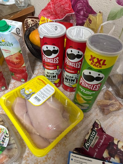 The image displays a collection of grocery items arranged on a light-colored, patterned tablecloth, indicative of an indoor setting within a residence in Ciorescu, Moldova. Central to the scene are three Pringles potato chip cans: two red cans of "Original" flavor, both featuring a "SCAN TO WIN A MONOPOLY TRIP TO NYC" promotion, and one green "XXL PACK SOUR CREAM & ONION" flavor can. To the left, a carton of "Naturalis Tomato Tomate" juice with a green screw cap is visible. In the foreground, a yellow plastic tray contains several raw chicken breasts, sealed under clear film, with a label indicating "CARNPRODLUX" and a weight of 900g, dated 05.02.20. A portion of a maroon-colored bag labeled "Alesto Selection Cashew & Cranberry" is present in the lower right corner. In the background, partially obscured, are a dark woven basket holding an orange fruit, a clear drinking glass, and various other indistinguishable packaged food items contained within plastic bags. No individuals are depicted, and no active interactions are occurring within the frame.