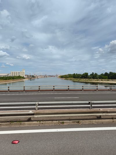 The image shows a view from a highway bridge over a river in Ottmarsheim, France. The river is wide and calm, reflecting the cloudy sky. On the left bank, there are industrial buildings with silos and smokestacks, suggesting a port or industrial area. On the right bank, there is a dense line of green trees. The foreground of the image shows the asphalt road of the bridge with white lane markings and a metal guardrail. A red reflective marker is visible on the road surface in the bottom left corner. The sky is filled with grey and white clouds, with some patches of blue sky visible. The overall scene suggests an industrial landscape along a waterway.