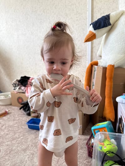 A young child, possibly a toddler, is standing in a room and holding a comb to their mouth. The child has light brown hair pulled into a messy ponytail with a red and yellow hair tie. They are wearing a white long-sleeved bodysuit with a pattern of brown teddy bears. The child's gaze is directed downward, and their left hand is holding the comb near their mouth, while their right hand is also near the comb.
The background shows a textured, light-colored wall. To the right of the child is a large, plush toy resembling a goose with orange legs and a dark blue and white head covering. Behind the goose and to the left of it, there are several cardboard boxes stacked. To the left of the child, there is a pile of clothing, including items in black, pink, and white. In the foreground, there is a blue plastic container and other toys are visible, including a green plastic toy resembling a frog in a clear plastic bin. The floor appears to be covered with a light-colored, textured material. The overall impression is that of a child playing in a room with toys and household items. The location is in the city of Vylygort, Russia.