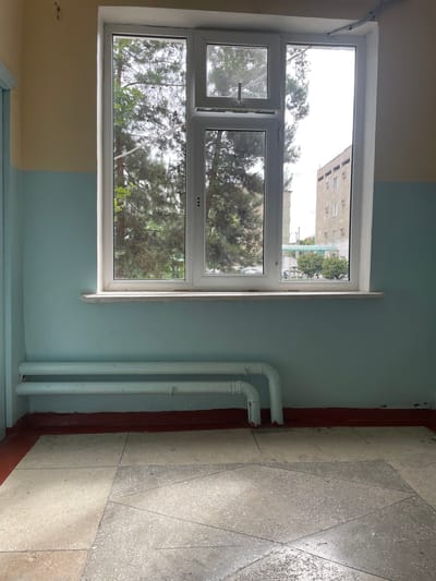 This is an outdoor, eye-level, medium shot of a white, multi-pane window in an interior hallway, possibly in Kyzyl-Kyya, Kyrgyzstan. The window, which appears to be made of PVC, has a light grey sill and is divided into four sections. Two large panes on the left and right, and two smaller panes stacked in the center. Through the window, lush green trees and a multi-story building are visible. The walls of the hallway are painted in two horizontal bands: a pale yellow on the top and a light blue on the bottom. Below the window, two white, horizontal pipes, likely part of a heating system, are mounted on the blue wall. A dark red baseboard runs along the wall above a grey, tiled floor. The overall impression is one of utilitarian architecture, possibly in a public building or residential complex.