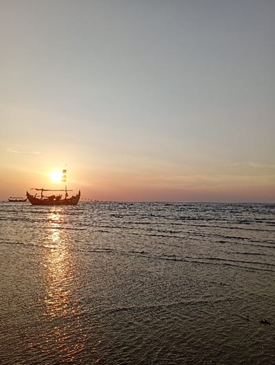 A wide shot captured at sunset or sunrise depicts a coastal scene in the city of Tritunggal, Indonesia. The sky occupies the upper two-thirds of the frame, displaying a gradient from light grey-blue overhead to warm orange and pink hues near the horizon. The sun, a prominent, bright orange disc, is positioned low on the left side of the horizon, casting a radiant, shimmering path of light across the water surface towards the foreground. Two traditional-style wooden boats are silhouetted against the illuminated sky. The primary boat, centered on the left, features a distinctive high bow and stern, and a visible mast-like structure. A smaller, simpler boat is present further to the left, behind the main vessel. The water, displaying gentle ripples, reflects the sun's light intensely on the left side. No individuals are visible.