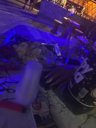 The image shows a dimly lit outdoor scene with a strong blue lighting cast. In the foreground, on a surface with a black and white patterned cloth, are a white plastic cup, a red cylindrical object, and a small box of tissues. To the right, a large dark bottle with a label containing the text "By Ia" is partially visible. Behind these items, a bag of potato chips is spread out, with some chips spilling out. Further back, there are wooden stools and tables, and a wire fence with lights behind it, suggesting an outdoor seating area of a bar or restaurant. The background is blurred, with figures of people visible, and the overall atmosphere is casual and appears to be nighttime. The image does not contain any information that would place it in Radievo, Bulgaria, or any other specific location.