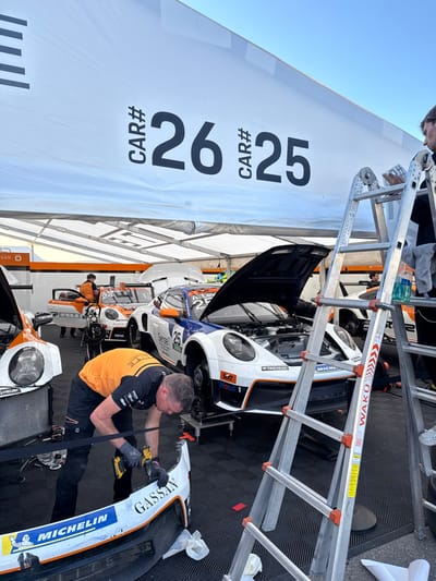 The image shows a pit stop area for race cars in Stavelot, Belgium. A mechanic wearing an orange shirt and black pants is working on the front end of a white race car with the number "25" on its side. The car is on a lift and is open at the front. The mechanic is using a drill. A second white race car, partially visible, is also on a lift behind the first car. Both cars have the word "Michelin" printed on their sides. The number "26" is displayed on a white banner overhead. There is a ladder beside the second car. The ground is black and checkered. The pit stop area is under a white tent. The background of the image is a cloudy sky.