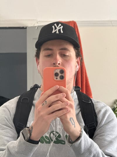 A young man takes a selfie in front of a mirror. He is wearing a grey sweatshirt with green lettering, a black baseball cap with a white New York Yankees logo, and a black crossbody bag. He has earphones in his ears and a silver watch on his left wrist. He is holding an orange iPhone in front of his face. His hands have tattoos, one being a number and the other an anchor. The location appears to be indoors, with a door in the background and an orange blanket. There is no visible information that the location is in Hitchin, United Kingdom.
