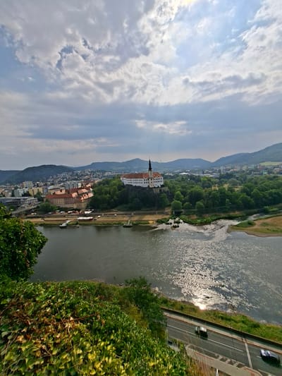 The image presents a wide-angle, elevated view of the city of Děčín, Czechia, with a focus on the Elbe River and a prominent castle complex. In the foreground, lush green vegetation, predominantly ivy, covers a steep embankment. Below this, the Elbe River flows from left to right, its surface reflecting the sunlight breaking through scattered clouds.

Along the riverbank opposite the vegetation, a road runs parallel to the water, with two cars visible. Beyond the road, the city of Děčín unfolds. Several multi-story buildings with red-tiled roofs are clustered together, some appearing older and more traditional, while others are modern concrete structures. A bridge spans a section of the river further into the cityscape.

Dominating the middle ground is the Děčín Castle, situated on a rocky outcrop amidst dense green trees. The castle features white walls and a tall, dark-spired tower. Beyond the castle and the immediate city, rolling hills covered in vegetation form the backdrop, their outlines softened by the distance and atmospheric conditions. The sky is partly cloudy, with patches of blue sky visible and sunlight intermittently illuminating the scene, creating bright reflections on the river. The overall impression is of a scenic riverfront city with historical architecture nestled within a natural landscape.
