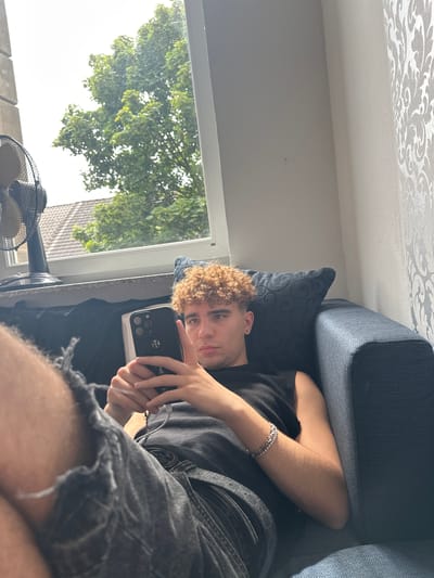A young man with curly blonde hair is lying on a sofa, holding a smartphone. He is wearing a black t-shirt and ripped denim shorts. A silver bracelet is visible on his left wrist. The sofa appears to be dark blue or grey. Behind him, a window shows a tree with green leaves, suggesting it is daytime and likely summer or spring. The room has a textured wallpaper on the ceiling with a damask pattern. A black fan is visible near the window. The overall impression is a casual, relaxed scene indoors. The image is taken from a low angle, looking up at the subject. The subject's gaze is directed towards the camera, and he is holding the phone in a way that suggests he might be taking a selfie or recording a video. The lighting indicates it is daytime, with natural light coming from the window. The background details suggest a domestic setting.