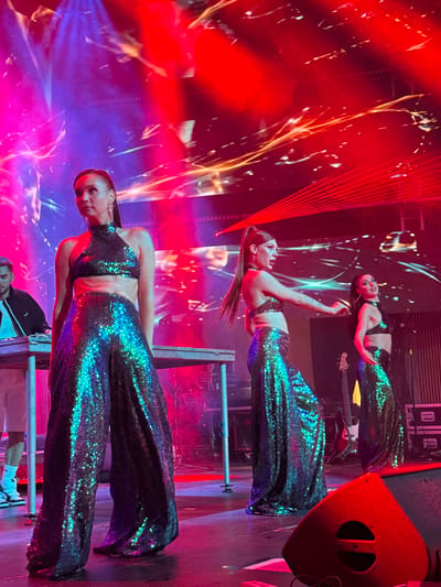 The image depicts a performance on a stage in Akhalubani, Georgia. Three women, dressed in shimmering, iridescent two-piece outfits consisting of halter tops and wide-legged pants, are the central focus. The woman in the foreground, on the left, stands facing forward, her hands at her sides. To her right, a second woman is captured mid-motion, her arm extended, looking to her left. A third woman, further to the right, is also in motion, looking towards her left. All three women have their hair pulled back.

The stage is illuminated by vibrant red and purple lighting, with streaks of light suggesting a dynamic visual display. Red laser beams are visible in the background, creating a dramatic effect. To the left of the stage, a man in a black jacket and white t-shirt is partially visible, seated behind a console with keyboards. Musical instruments, including a guitar, are placed on the stage behind the performers. Stage equipment, such as speakers and lighting structures, are also present. The overall environment suggests a live music or performance event.