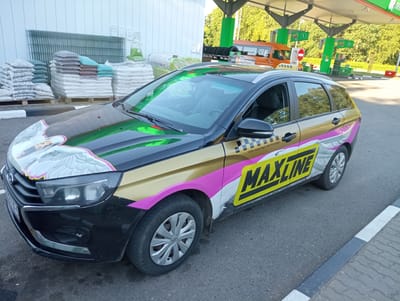 A black station wagon with a gold, pink, and white racing stripe design and "MAXLINE" text is parked on an asphalt surface with a gray and white curb. The car has a graphic of a white bird with pink and yellow accents on its hood. The background shows a light-colored building, stacks of bagged materials on pallets, a gas station with green and white pumps, and an orange van. The image appears to be taken on a sunny day.