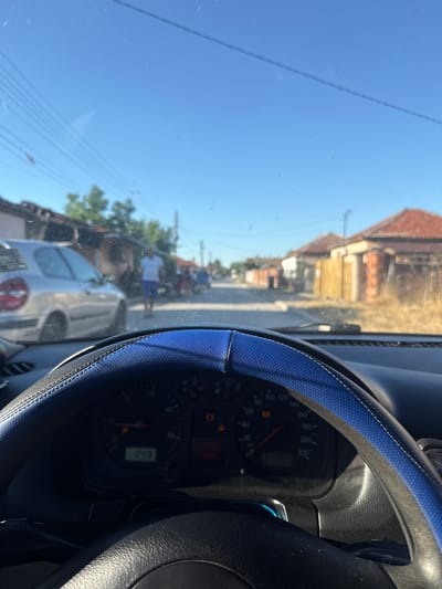 The image is taken from inside a car, looking forward through the windshield. The car's dashboard and a portion of the steering wheel are visible in the foreground. The steering wheel is covered with a blue and black fabric. To the left of the steering wheel, the speedometer shows a reading of 24, indicating a speed of 24 km/h. Several indicator lights are illuminated on the dashboard.
Outside the car, the scene depicts a street in a residential area. The sky is clear and blue. On the left side of the street, a silver car is parked. Further down the street, a person wearing a white t-shirt and blue shorts is walking. Houses with red-tiled roofs line both sides of the street. Some of the houses appear to be older or in disrepair, with visible brickwork and wooden fences. There are trees and dry vegetation along the sides of the road. In the distance, other vehicles, including what appears to be a scooter or motorcycle, are also visible on the street. The environment suggests a sunny day in a village or a less developed part of a city. This scene is consistent with a street in Vinitsa, Bulgaria, given the architectural style of the houses and the general atmosphere.