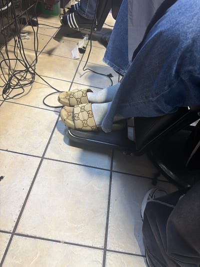 An overhead shot captures two individuals' lower bodies and a cluttered tiled floor in an indoor setting, likely a salon or barbershop in Blouberg, South Africa.
On the left, a person is wearing blue denim jeans, light grey socks, and tan slip-on sandals adorned with the interlocking "Gucci" G logo pattern. Their left foot rests on a black footrest or part of a chair. Partially visible on the right, another person wears dark grey or black pants and a white athletic shoe featuring black details and three prominent stripes on the side, characteristic of Adidas.
The floor is composed of light-colored square tiles with dark grey grout lines. Numerous tangled black electrical cords are strewn across the floor. Also visible are small pieces of white disposable material, possibly wipes or tissues, and dark debris, likely hair clippings, scattered on the tiles. A green container is partially visible on the far left. The scene appears to be active during the daytime.