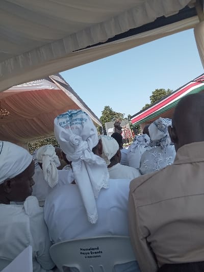 People wearing white clothing are kneeling, possibly praying, under a tent. A Kenyan flag is visible in the background. Some people are wearing head coverings. A white plastic chair has the words "Homeland Hoya Events" printed on it. The overall impression is of a religious gathering or ceremony.