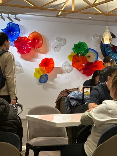 The image depicts an interior space, identified as a lounge or waiting area within Taiwan Taoyuan International Airport in Taoyuan City, Taiwan. A white wall dominates the background, featuring an extensive installation of vibrantly colored, organically shaped glass discs, including shades of blue, red, orange, green, yellow, and clear. These discs are mounted in clusters and illuminated by recessed ceiling spotlights, generating pronounced shadows on the wall. Wooden ceiling beams form an overhead grid.

To the left, a male figure wearing a tan outer layer and dark pants, with a dark backpack visible, stands in partial profile, facing the glass art. In the midground and foreground right, multiple individuals are partially visible, seated around white rectangular tables with grey upholstered chairs. One subject wears a light grey hooded garment, with an arm resting on a table. Other visible subjects include a person in a dark blue shirt, and a dark-haired individual in a dark polka-dotted top. Luggage, including a dark wheeled bag, is situated near the tables, and a brown jacket is draped over a chair. A book with a light green cover and Chinese script is present on a table surface.