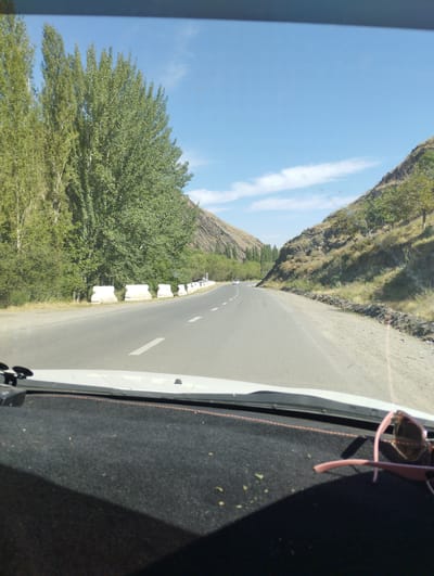 The image depicts a view from inside a vehicle traveling on a paved road through a mountainous landscape. Tall, leafy trees line the left side of the road, with a rocky, sparsely vegetated hillside on the right. The road has white lane markings and concrete barriers are visible along the left shoulder. A white car is seen in the distance ahead. The sky is blue with scattered white clouds. The dashboard of the car is visible in the foreground, with a pair of pink sunglasses resting on it. The scene appears to be set in a rural, natural environment, possibly in the vicinity of Karangisay, Uzbekistan, given the landscape and vegetation.
