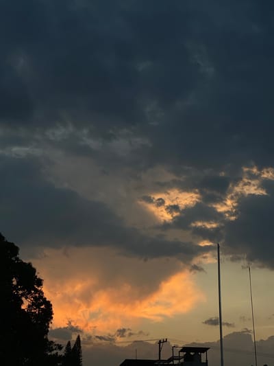 The image shows a cloudy sky in Santa María del Tule, Mexico. Dark, heavy clouds dominate the left side of the frame. On the right, clouds are illuminated with a warm orange and yellow light, suggesting a sunset or sunrise. Silhouetted trees are visible at the bottom, and utility poles or wires run vertically through the right side of the frame, indicating an urban or suburban environment. The image is rotated 90 degrees clockwise.